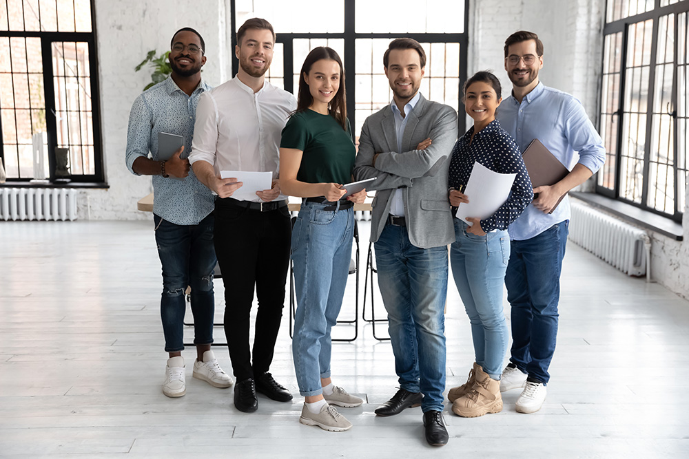 Portrait of happy millennial diverse professional team in loft office space. Group of multi ethnic employees gathering for corporate meeting and teamwork, looking at camera, smiling. Full length